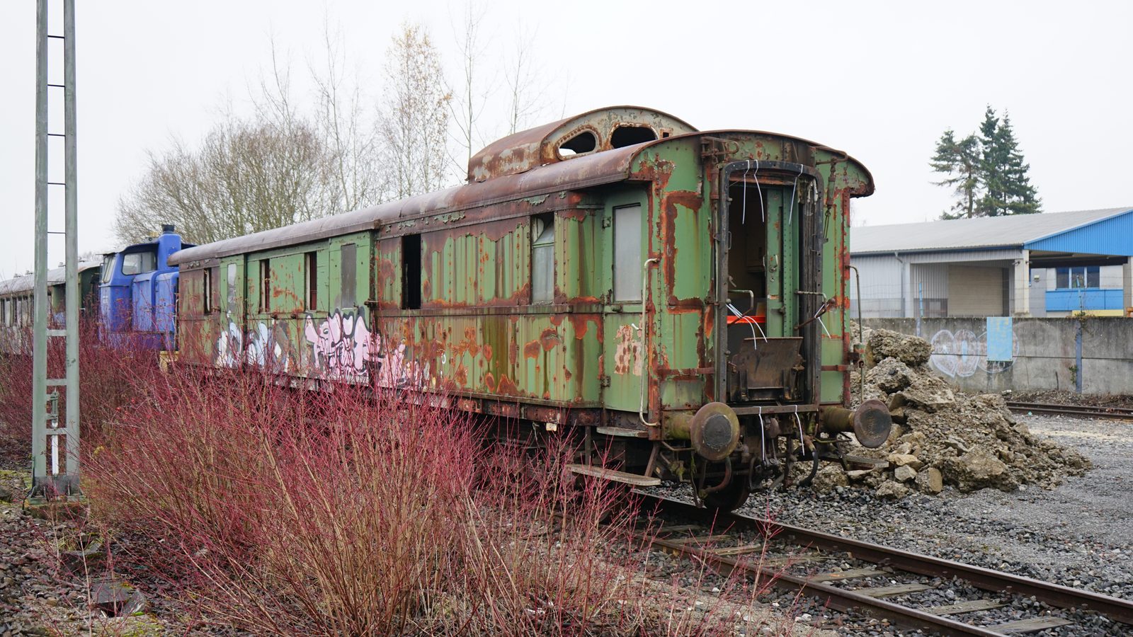 Grüner historischer Reisezug-/Gepäckwagen mit Rost und Graffiti auf einem Abstellgleis, blaue Rangierlok im Hintergrund (Winter).